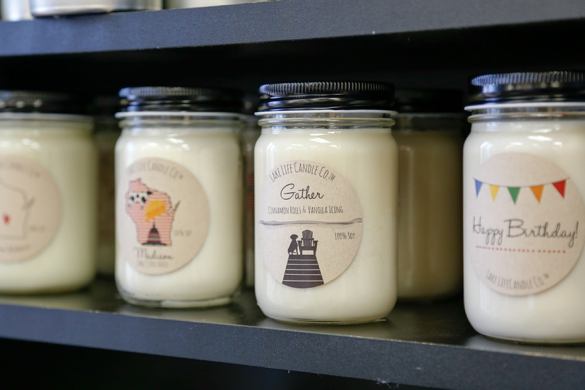 Soy candles on a shelf, featuring labels with playful designs and scents like "Gather" and "Happy Birthday."