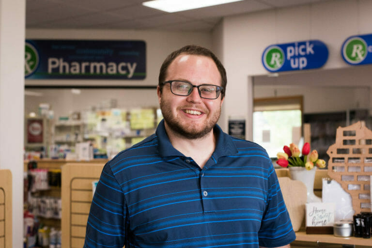 Smiling man in a blue striped polo stands in a pharmacy, with shelves of products and a vase of tulips in the background.