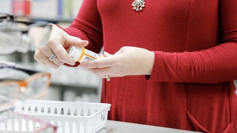 A person in a red shirt holds a prescription bottle, surrounded by eyeglasses and a white basket on a counter.