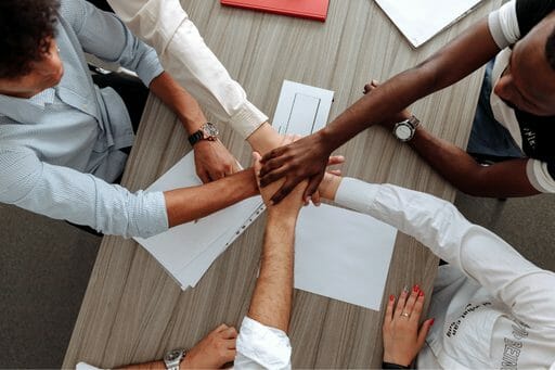 Hands of diverse individuals stacked together on a table, symbolizing teamwork and collaboration, with papers scattered around.