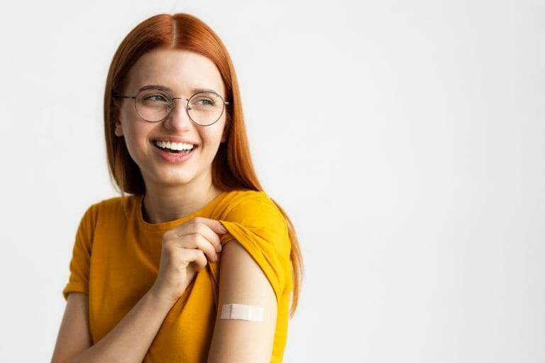 Smiling young woman in a yellow shirt shows a bandage on her arm against a plain white background.