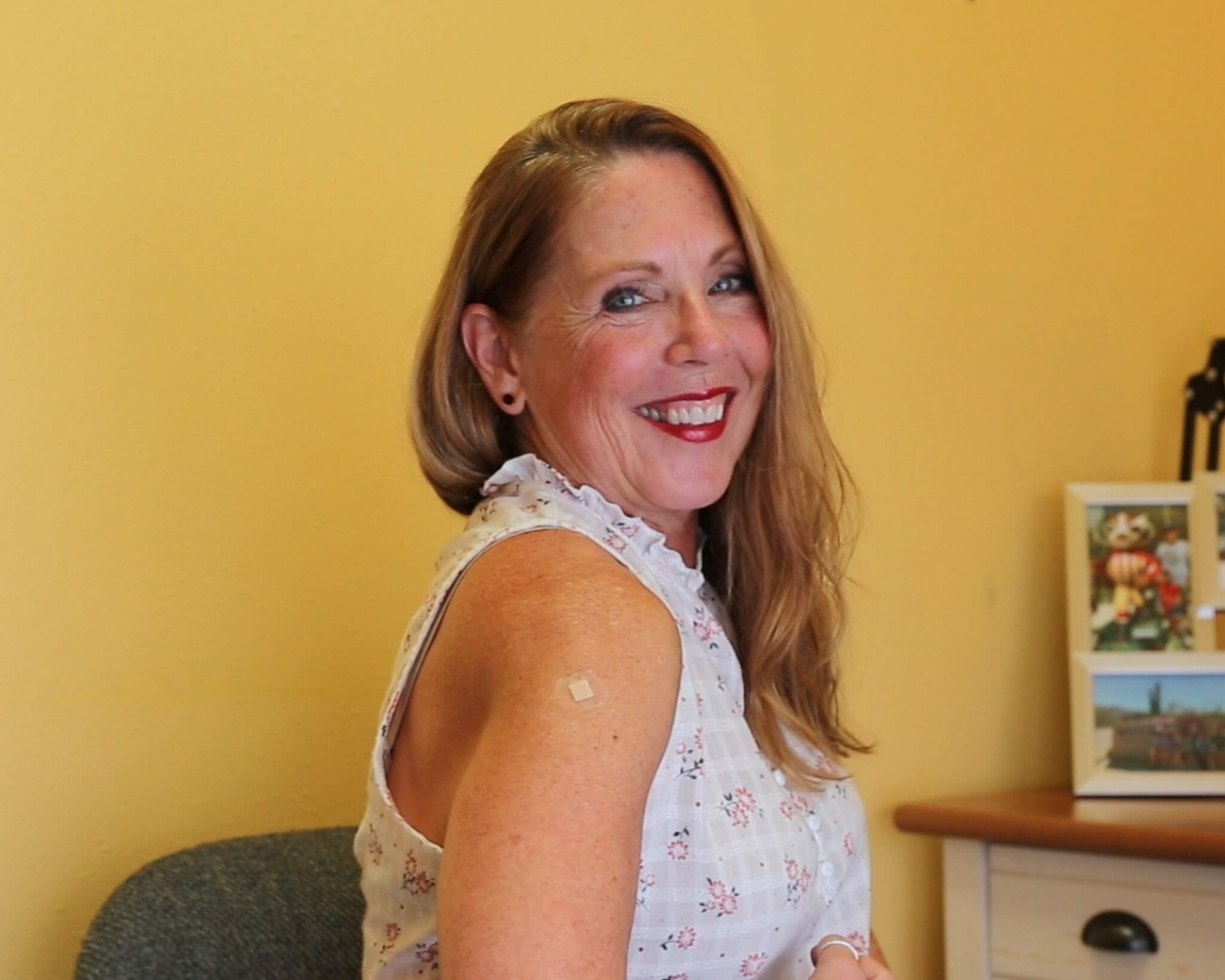 Woman with long hair smiles over her shoulder, showing a bandage on her arm against a yellow wall and a desk with photos.