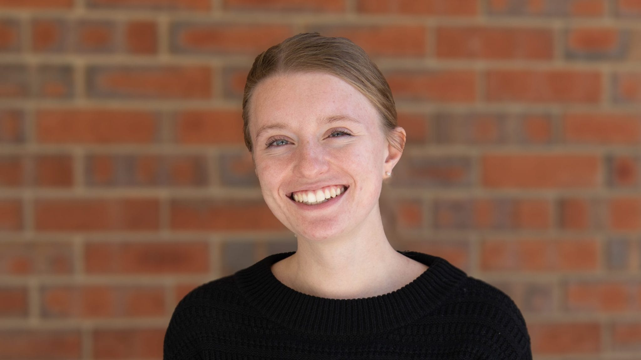 Smiling young woman in a black sweater against a brick wall background.