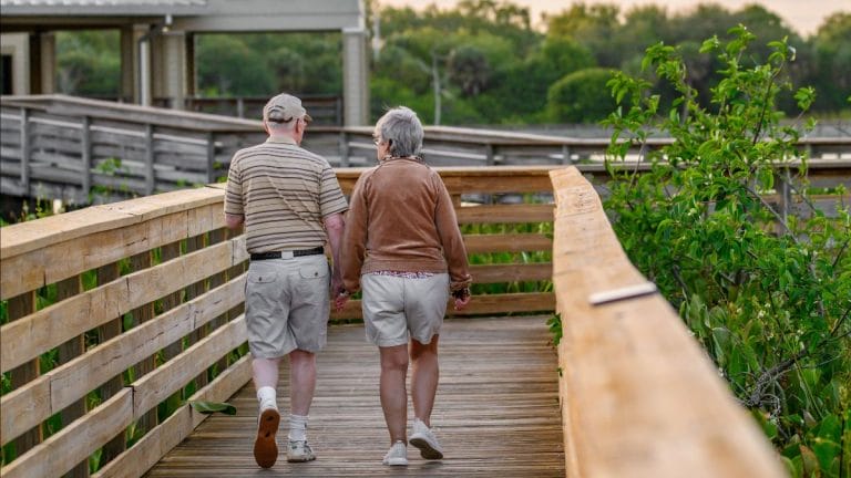 An elderly couple walks hand in hand along a wooden boardwalk surrounded by greenery at sunset.