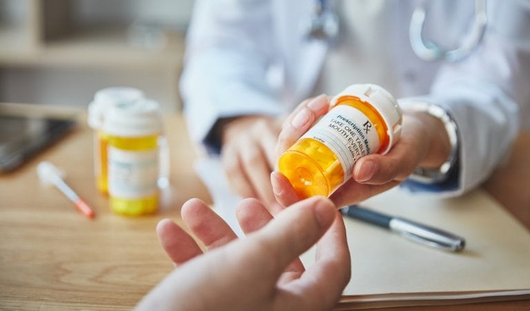 A healthcare professional hands a prescription bottle to a patient, with additional medication bottles visible in the background.