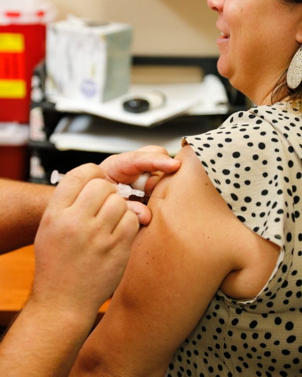 A healthcare professional administers a vaccine to a woman’s upper arm, with a polka dot shirt and a smiling expression.