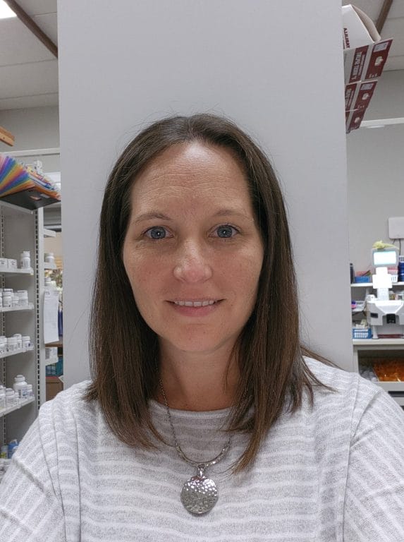 Smiling woman with long brown hair, wearing a striped shirt and a silver pendant, stands in a pharmacy setting.
