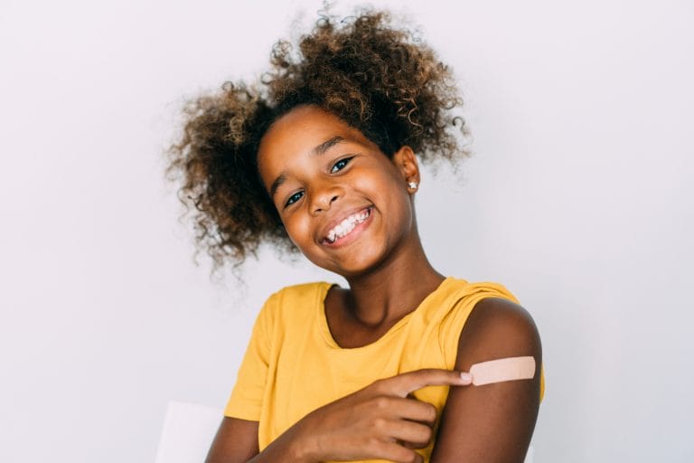 Smiling girl with curly hair in a yellow shirt points to a bandage on her arm against a light background.