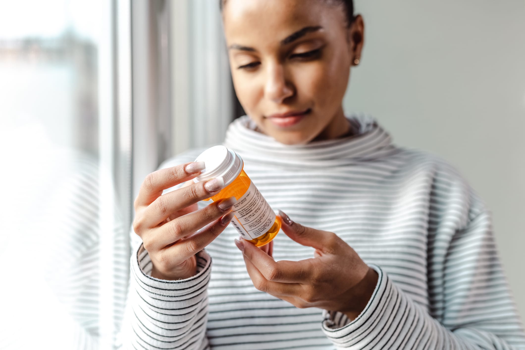 A woman in a striped sweater examines a prescription bottle, focusing intently on the label. Natural light filters in.