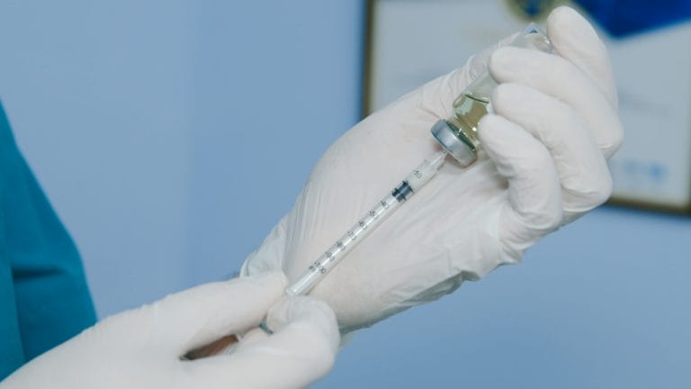 A gloved hand holds a syringe filled with liquid, poised above a vial against a soft blue background.