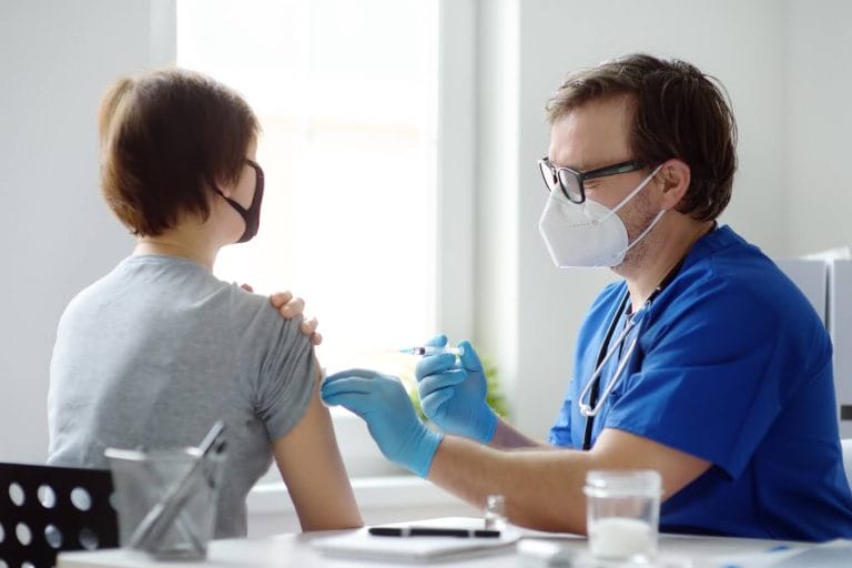 A healthcare worker administers a vaccine to a masked patient in a bright, clinical setting.