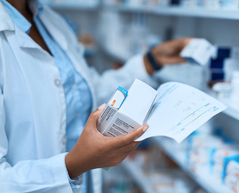 A pharmacist examines a medication box while holding a prescription in a pharmacy filled with various products.