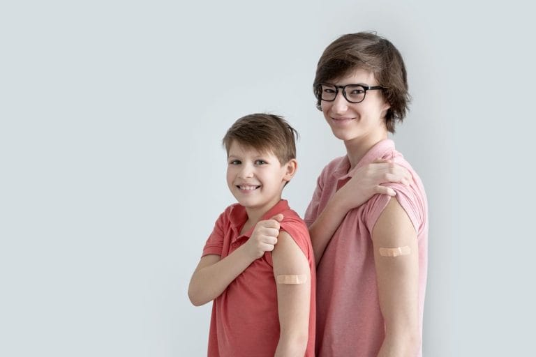 Two boys in matching pink shirts smile, showing band-aids on their arms against a light gray background.