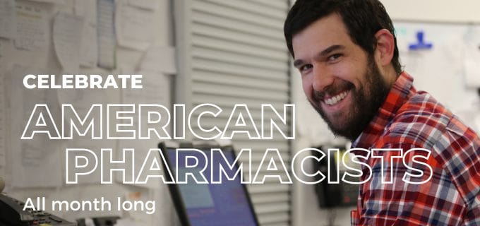 Smiling pharmacist in a checkered shirt at a workstation, surrounded by paperwork, promoting American Pharmacists Month.