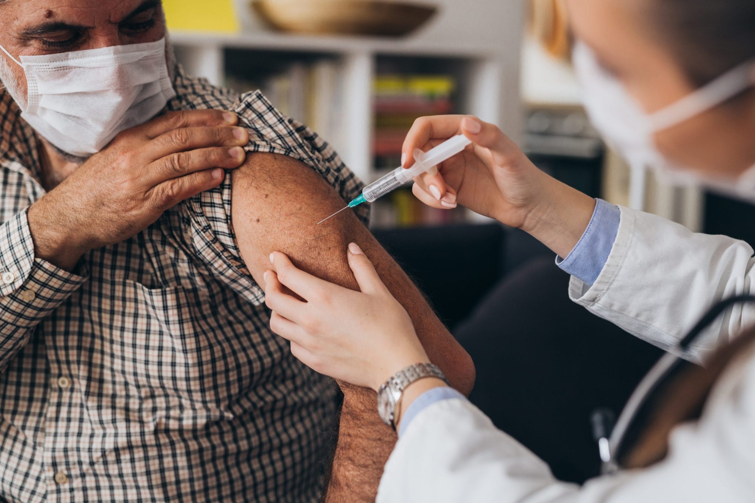 A healthcare professional administers a vaccine to an elderly man, both wearing masks in a cozy indoor setting.