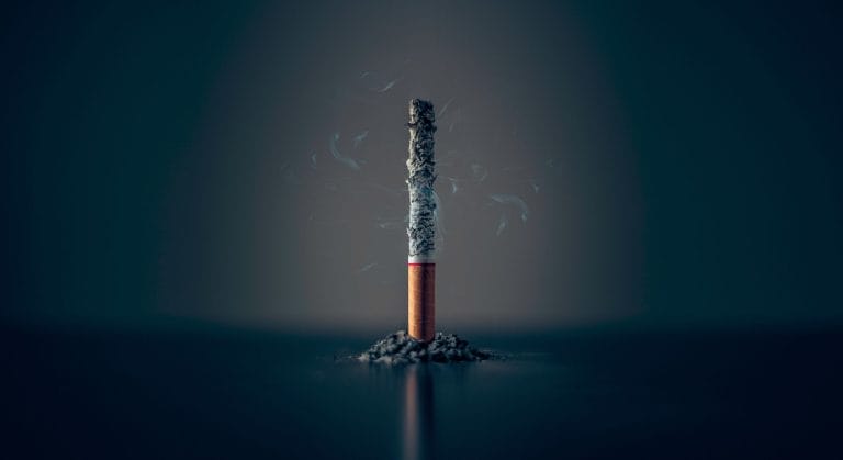 A lit cigarette stands upright on a mound of ash, with wisps of smoke curling upwards against a dark background.