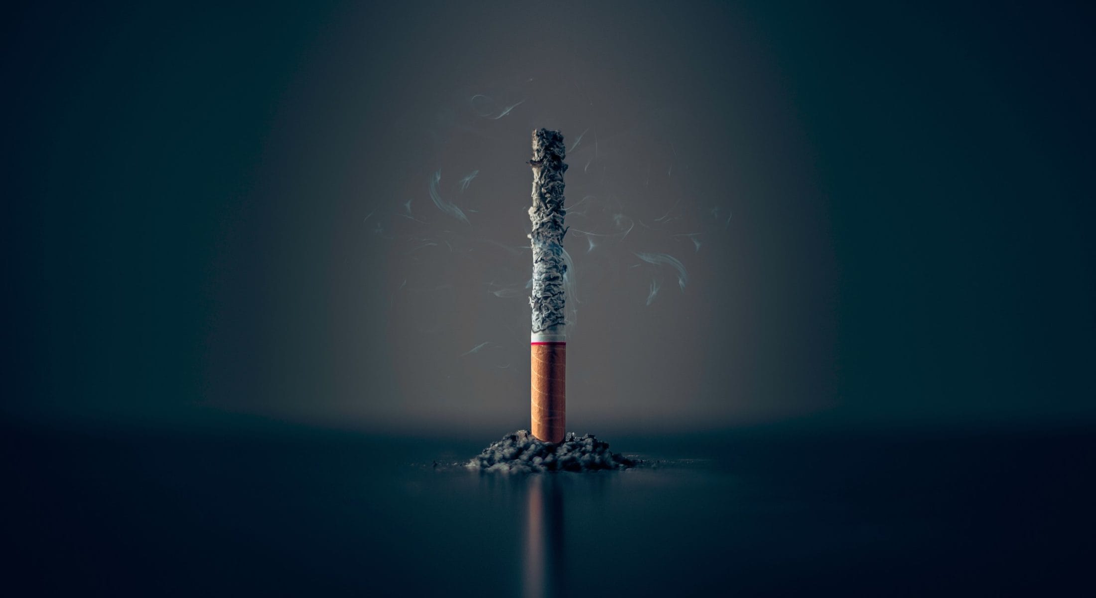 A lit cigarette stands upright on a mound of ash, with wisps of smoke curling upwards against a dark background.