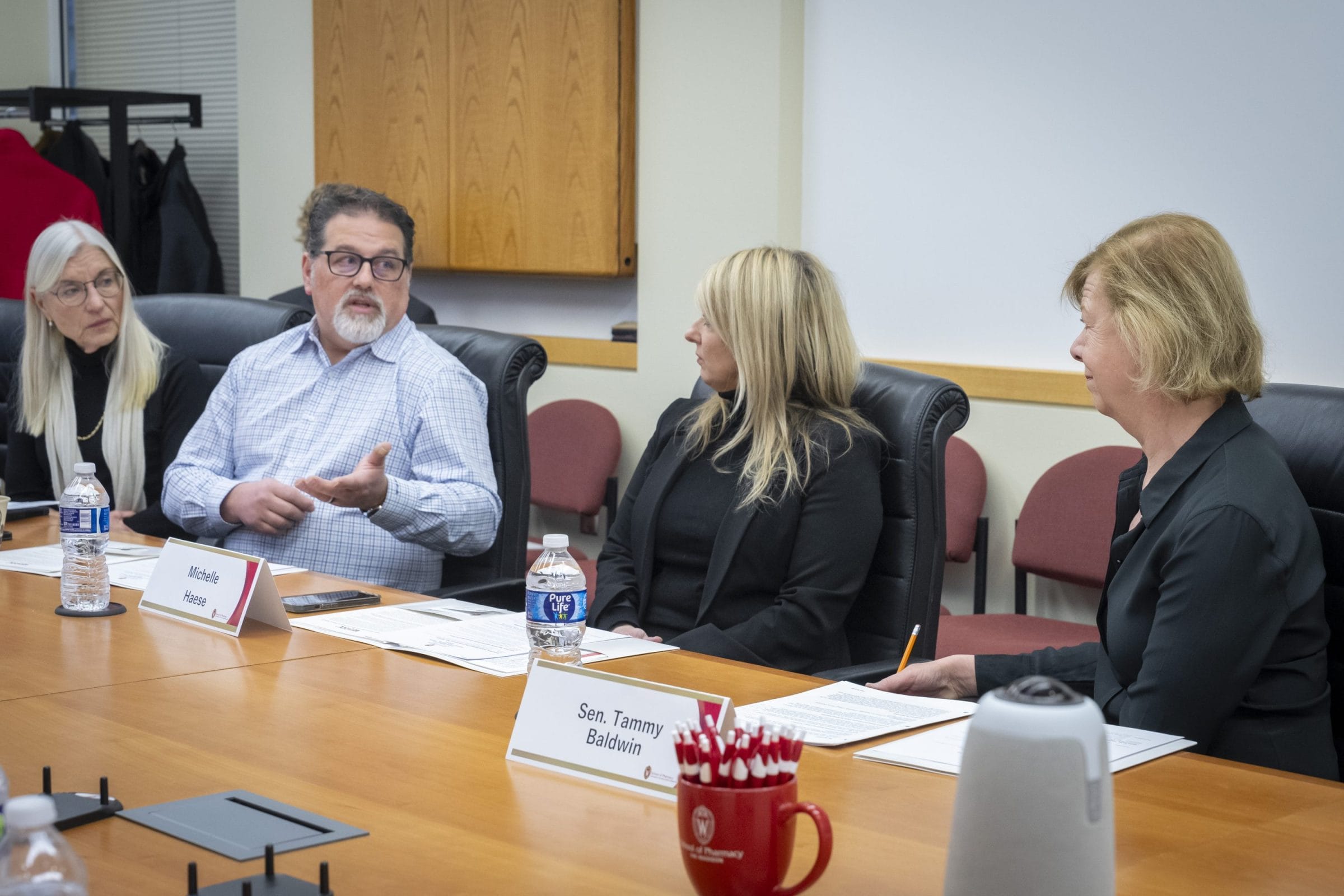 A group discussion at a conference table with four participants, water bottles, and documents in a professional setting.