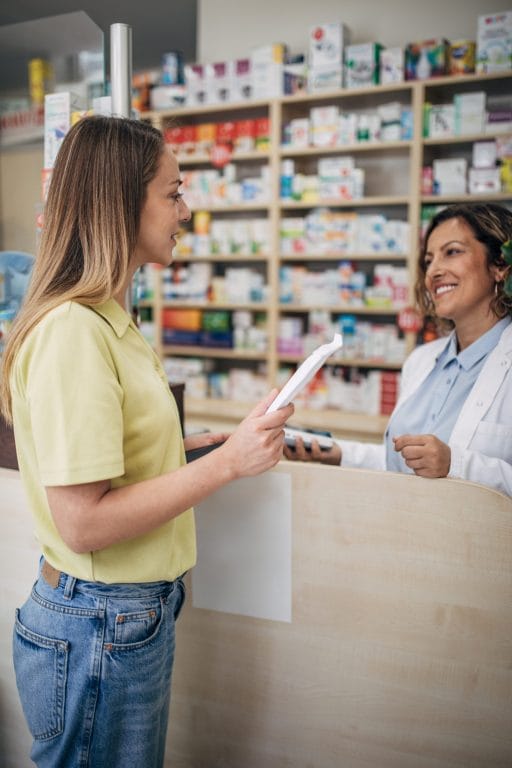 A woman in a yellow shirt interacts with a smiling pharmacist at a pharmacy counter, surrounded by medication shelves.