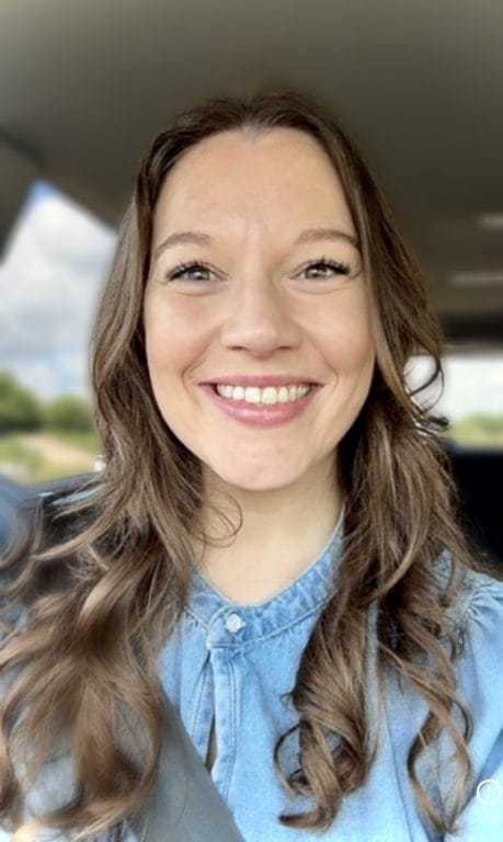 Smiling woman with long, wavy hair wearing a denim shirt, seated in a car with a blurred outdoor background.